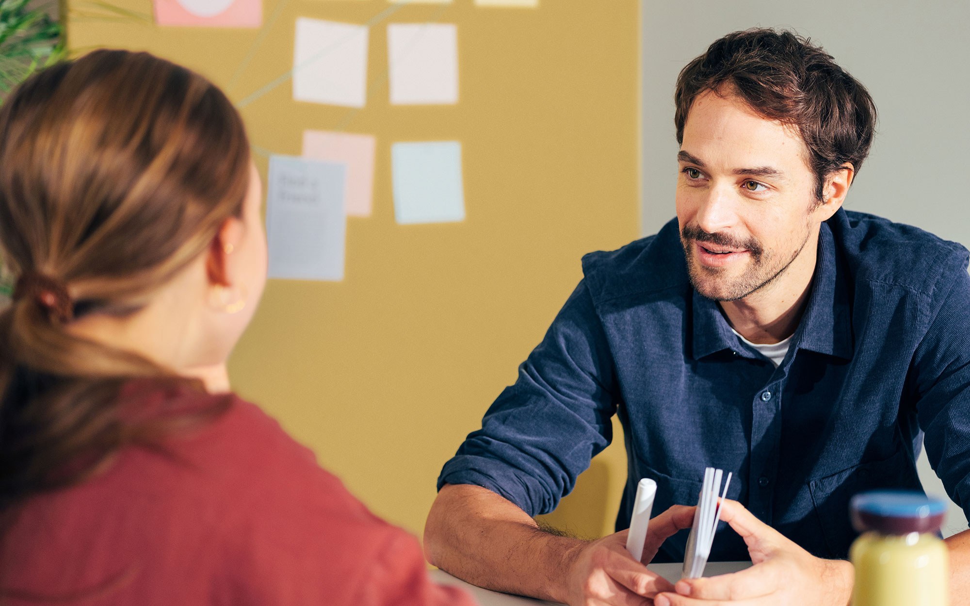 Eine Frau und ein Mann sitzen einander gegenüber bei einem Besprechungstisch und tauschen sich aus. Im Hintergrund ist eine Pinnwand mit Post-its zu sehen.
